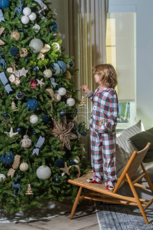 Foto de Feliz Navidad. Longitud completa de niño feliz decorando el árbol de Navidad en casa. Alegre niño lindo árbol de navidad decorado. Niños con pelotas de Navidad y juguetes cerca del árbol en la habitación - Imagen libre de derechos