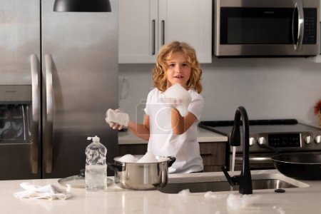 Photo for Kid boy washing dishes in the kitchen interior. Child with sponge with dish washing liquid is doing the dishes at home kitchen by using wash sponge and dishwashing - Royalty Free Image