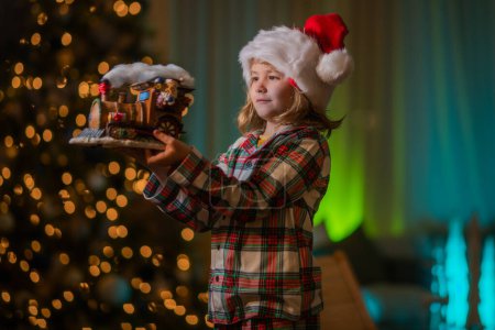 Foto de El chico se está divirtiendo cerca del árbol de Navidad en casa por la noche. Feliz Navidad y Feliz Año Nuevo. Niño celebrando la víspera de Navidad cerca de naght brillante guirnalda de Navidad - Imagen libre de derechos