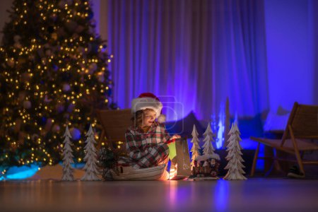 Téléchargez les photos : Cadeaux d'ouverture pour enfants la veille de Noël. Éclairage dans la nuit de Noël magique à la maison confortable. Enfant mignon le soir de Noël à la maison. Joyeux Noël - en image libre de droit