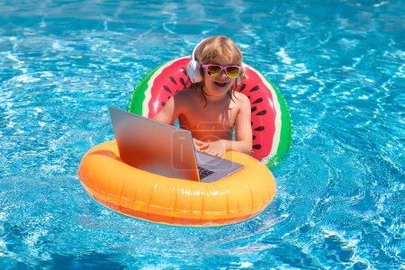 Téléchargez les photos : Utilisation d'un ordinateur portable dans la piscine. Les affaires d'été. Kid télécommande de travail sur ordinateur portable dans la piscine. Petit homme d'affaires travaillant en ligne sur ordinateur portable dans l'eau de piscine d'été - en image libre de droit