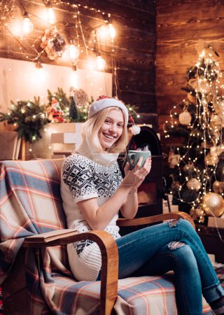 Photo for Santa girl in cozy white sweater sitting at sofa and drinking hot tea, cacao, coffee at wonderful christmas decorated background. Merry christmas and happy new year. Christmas gifts - Royalty Free Image