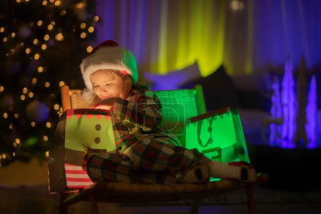 Téléchargez les photos : Enfant avec cadeau cadeau avec lumière magique. Éclairage cadeau sac cadeau. Enfant en chapeau de Père Noël sur le devant de l'arbre de Noël de nuit arrière-plan de la maison la veille de Noël - en image libre de droit