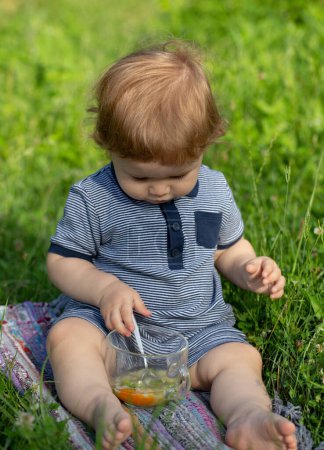 Téléchargez les photos : Bébé garçon manger de la purée de légumes en plein air sur herbe verte - en image libre de droit