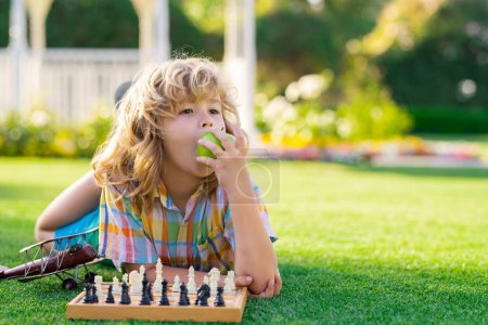 Téléchargez les photos : École d'échecs en plein air. L'enfant pense ou planifie le jeu d'échecs, pose sur l'herbe, manger des pommes dans le parc d'été. Des écoliers intelligents, intelligents et intelligents - en image libre de droit