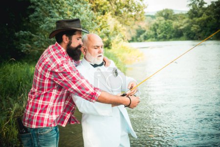 Foto de Retrato de un alegre hombre mayor pescando. Abuelo e hijo pescadores. Joven y un viejo pescando para spinnings en el río o el lago - Imagen libre de derechos