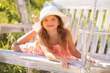 Foto de Niña feliz riéndose y balanceándose en un columpio en el jardín de verano. Niña divirtiéndose en un columpio al aire libre. Retrato de una hermosa niña con sombrero soñando - Imagen libre de derechos