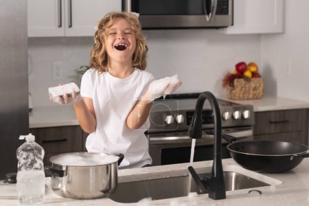 Téléchargez les photos : Portrait d'un garçon de 7-8 ans qui lave la vaisselle à la maison. Enfant femme de ménage laver la vaisselle sur de l'eau savonneuse. mignon drôle garçon lavage vaisselle dans cuisine - en image libre de droit