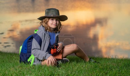 Téléchargez les photos : Joli gamin blond avec des jumelles portant un chapeau explorateur et un sac à dos sur la nature. Enfant explorateur randonnée et aventure - en image libre de droit