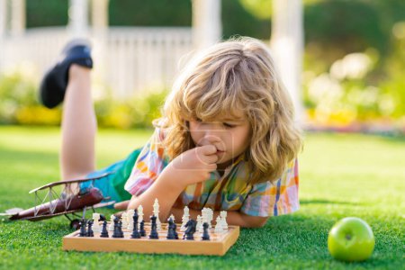 Téléchargez les photos : Enfant concentré développant une stratégie d'échecs, jouant au jeu de plateau dans la cour arrière, s'allongeant sur l'herbe - en image libre de droit