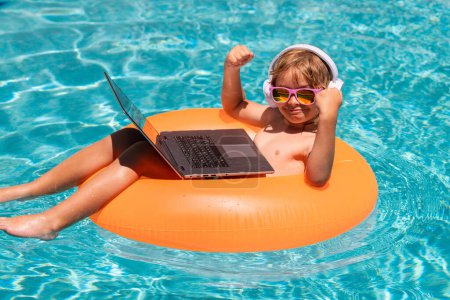 Téléchargez les photos : Les affaires d'été. Joyeux petit garçon apprenant ordinateur portable en ligne. Enfant se détendre sur anneau flottant en utilisant un ordinateur dans la piscine - en image libre de droit