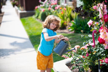 Téléchargez les photos : Enfant mignon garçon arrosage des plantes et des roses avec arrosoir dans le jardin. Enfant en t-shirt d'été, souriant et amusant. Activités avec les enfants en plein air - en image libre de droit
