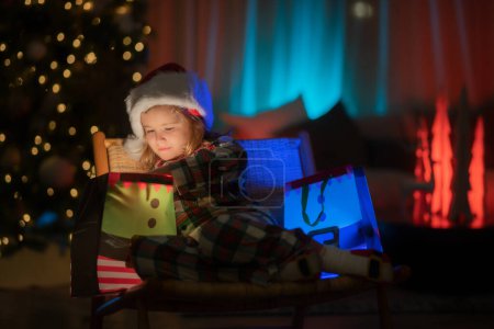 Foto de Niño con regalo presente con luz mágica. Iluminación regalo bolsa. Niño en pijama de Navidad disfrutando de la noche de vacaciones en casa - Imagen libre de derechos