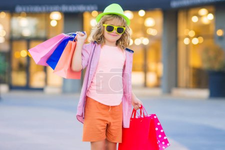 Photo for Child in fashion clothes goes shopping. Kid with shopping packages outdoor. Shopper child with shopping bag walking on street and carrying shopping bags - Royalty Free Image