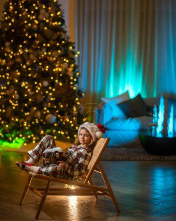 Téléchargez les photos : Joyeux enfant en pyjama près du sapin de Noël à la maison avec arbre de Noël traditionnel. Cozy chaude maison de soirée d'hiver - en image libre de droit