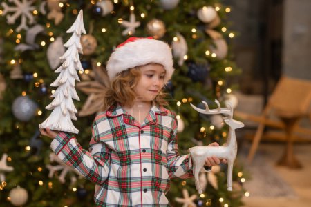 Foto de El niño feliz sostiene una bola de Navidad. El muchacho se alegra en la fiesta, la Navidad, el Año Nuevo. El chico se levanta contra el árbol de Navidad. Juguetes Navidad - Imagen libre de derechos