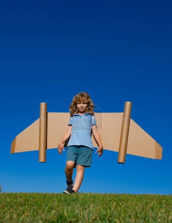 Foto de Niño feliz jugando con alas de juguete contra el fondo del cielo de verano. Éxito infantil, líder y concepto ganador - Imagen libre de derechos