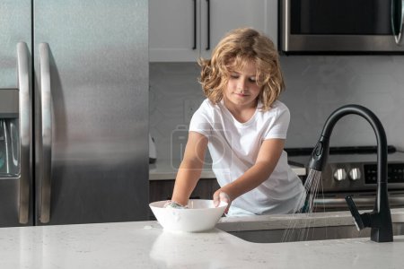 Photo for Kid boy washing dishes in the kitchen interior. Child helping his parents with housework - Royalty Free Image