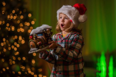 Téléchargez les photos : Enfant célébrant Noël à la maison. Enfant en santa chapeau sur le devant de l'arbre de Noël de nuit arrière-plan de la maison la veille de Noël. Joyeux enfant en pyjama de Noël à carreaux profitant d'une soirée de vacances à la maison - en image libre de droit