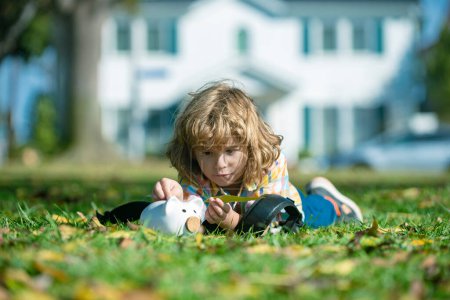 Photo for Portrait of a little boy putting money on a moneybox. Child saving money in a piggybank on backyard outdoor - Royalty Free Image