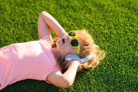 Téléchargez les photos : Enfant écoutant de la musique dans les écouteurs. Enfant mignon en lunettes de soleil d'été profitant de la musique extérieure - en image libre de droit