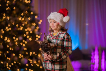 Téléchargez les photos : Le gamin s'amuse près du sapin de Noël la nuit. Enfant en pyjama de Noël profitant soirée de vacances d'hiver à la maison près de l'arbre de Noël de nuit - en image libre de droit