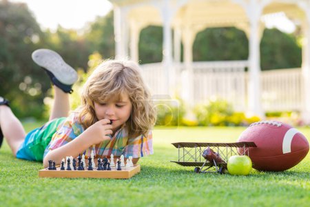 Téléchargez les photos : Jeux d'été et activités de plein air pour les enfants. Enfant jouant au jeu d'échecs dans le jardin de printemps, allongé sur l'herbe. Un gamin concentré joue aux échecs. Enfant jouer jeu de société en plein air - en image libre de droit