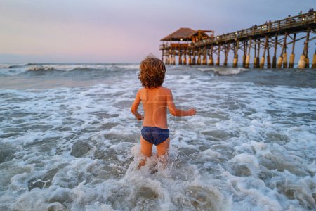 Téléchargez les photos : Enfant jouant en plein air sauter dans l'eau pendant les vacances d'été sur l'île de plage tropicale. Joyeux enfant jouant dans la mer. Océan bleu avec de grosses griffes blanches sur le fond - en image libre de droit
