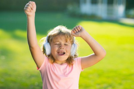 Foto de Cantando y bailando niños. Niño feliz disfruta escuchando música en los auriculares sobre el fondo de hierba verde. Divertido niño en auriculares escuchando música en el parque de verano o patio trasero al aire libre - Imagen libre de derechos