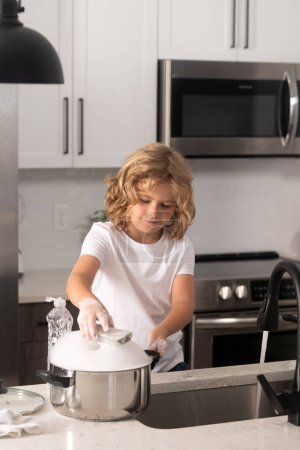 Téléchargez les photos : Enfant garçon laver la vaisselle dans l'évier de la cuisine. Nettoyage vaisselle pendant les travaux ménagers - en image libre de droit