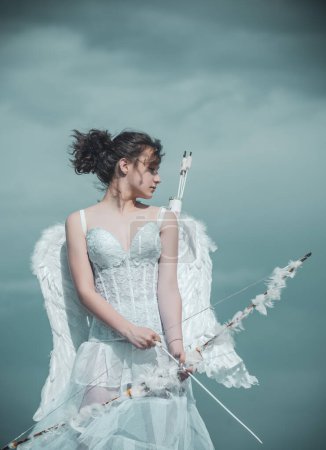 Foto de Valentin adolescente con flecha de arco listo para disparar. Angel adolescente en ángeles blancos vestido con alas de plumas en el cielo. Día de San Valentín - Imagen libre de derechos