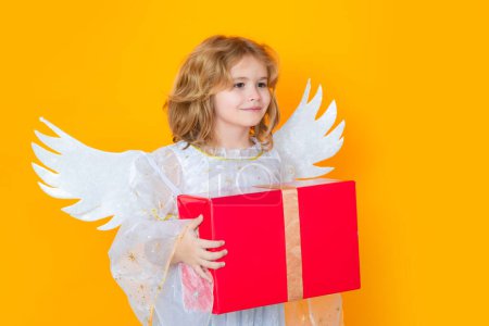 Photo for Cute blonde kid angel with gift box present. Cute angel kid, studio portrait. Blonde curly little angel child with angels wings, isolated background - Royalty Free Image