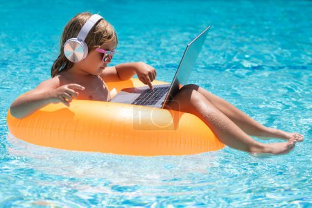 Foto de Trabajo en línea y relajarse en la piscina. Niño trabajando en el ordenador portátil en la piscina junto a la piscina. Tecnología en línea de verano - Imagen libre de derechos