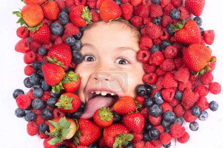 Téléchargez les photos : Des fruits drôles. Les enfants font face avec des fruits et des baies. Fraise d'été, myrtille, framboise, fond de mûre. Vue du dessus photo du visage de l'enfant en arrière-plan de baies. Enfants en bonne santé manger - en image libre de droit