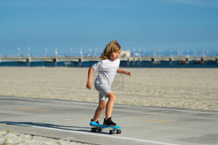 Foto de Young boy on his skateboard striking and practices his skill at the skate park. Happy child having fun on summer holidays. Pretty little kid learning to skateboard outdoors on beautiful summer day - Imagen libre de derechos