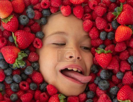 Téléchargez les photos : Funny fruits. Kids face with close-up berry. Berries mix of strawberry, blueberry, raspberry, blackberry. Assorted mix of strawberry, blueberry, raspberry, blackberry with background near child face - en image libre de droit
