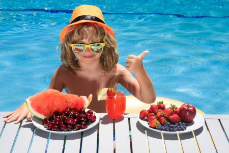 Foto de Healthy food. Outdoor leisure activity with kids by swimming pool. Summertime. Kid with fruits and juice smoothie cocktail in summer pool. Child with thumbs up on summer vacation. Summer fruits - Imagen libre de derechos