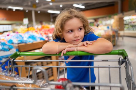 Photo pour Kid with shopping cart at grocery store. Kid is choosing fresh vegetables and fruits in the store. Child buying food in grocery supermarket. Buying in grocery store. Groceries in the supermarket - image libre de droit