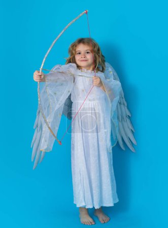 Foto de Angel shoots a love arrow from a bow on Valentines Day. Kid wearing angel costume white dress and feather wings. Innocent child. Little angel on isolated studio background - Imagen libre de derechos