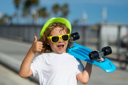 Foto de Excited child with thumb up, street portrait close up. Kid with skateboard. Child hold skate board. Healthy sport and activity for school kids in summer. Sports fun - Imagen libre de derechos