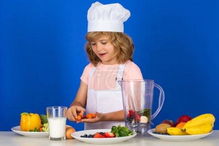 Téléchargez les photos : Portrait of chef child in cook hat hold carrot. Cooking at home, kid boy preparing food from vegetable and fruits. Healthy eating - en image libre de droit