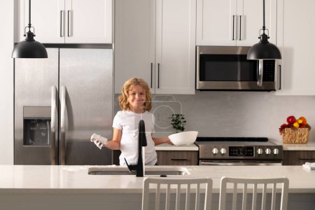Foto de Child housekeeper washing the dishes on soapy water. Cute Funny boy washing dishes in kitchen. Dishwashing liquid bottle on kitchen sink and clean plates - Imagen libre de derechos
