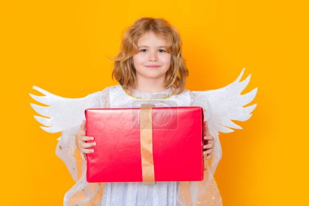 Téléchargez les photos : Cute blonde kid angel with gift box present. Little angel. Portrait of cute kid with angel wings isolated on studio background. Little angel, valentines day. Angelic kids - en image libre de droit