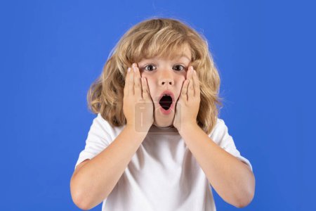 Téléchargez les photos : Shocked kid boy keeping hands near cheek with open mouth on blue isolated background. Surprised face, excited emotions of child. Shock, omg and wow expression - en image libre de droit