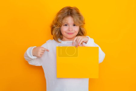 Photo for Cute child showing index finger on sheet of paper, isolated on yellow background. Portrait of a kid holding a blank placard, poster - Royalty Free Image