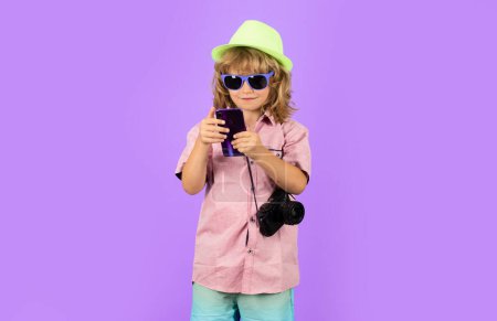 Téléchargez les photos : Child holding mobile phone and camera, traveling photographer. Fashion boy with summer hat, shirt and sunglasses. Travel and tourism concept - en image libre de droit