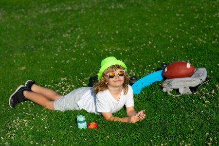 Téléchargez les photos : Child relax at the summer park. Kid boy lying on green grass outdoor. Outdoor portrait of cute kid wearing sunglasses, summer hat, t-shit and short - en image libre de droit