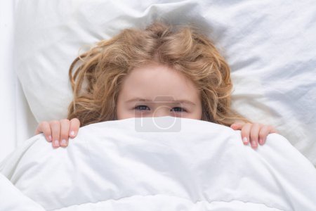 Téléchargez les photos : Kid under covers, face cover with blanket. Child wakes up in the morning in the bedroom. Cute little boy waking up in bed - en image libre de droit