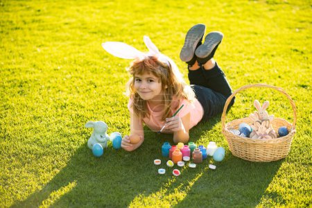 Foto de Child laying on grass in park wit easter eggs. Child boy with easter eggs and bunny ears laying on grass painting eggs - Imagen libre de derechos