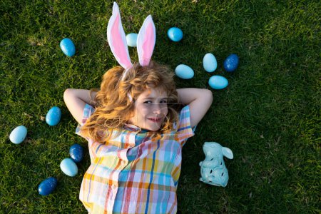Foto de Conejito de Pascua niño cazando huevos al aire libre Feliz día de Pascua, Niño que pone en la hierba en el parque. Vista superior cara de los niños con orejas de conejo - Imagen libre de derechos